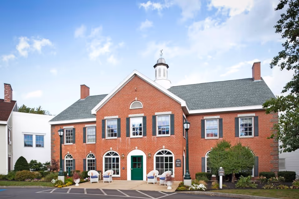 Front exterior view of a two-story brick building with green shutters and a green door, featuring a small cupola on the roof. There are four white chairs with cushions arranged outside near the entrance, along with some potted plants and landscaping. The sky is partly cloudy with blue patches.