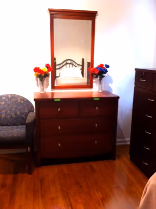 Wooden dresser with a mirror in a bedroom, flanked by vases of flowers, a patterned armchair to the left and a tall drawer unit to the right on hardwood floors.