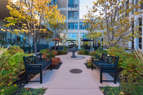 Outdoor courtyard area at Mt. Carmel Rehabilitation and Nursing Center featuring a paved walkway with two black benches facing each other, surrounded by landscaped greenery and trees with autumn foliage. In the background, there are tables with umbrellas and a multi-story building with large windows.