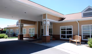 Exterior view of the entrance to Brookdale Boise Parkcenter facility showing a covered drop-off area with columns, benches, windows, and surrounding greenery under a clear sky.
