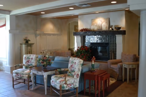 Cozy interior seating area with floral armchairs, a sofa, side tables, and a fireplace with a decorative mantle.