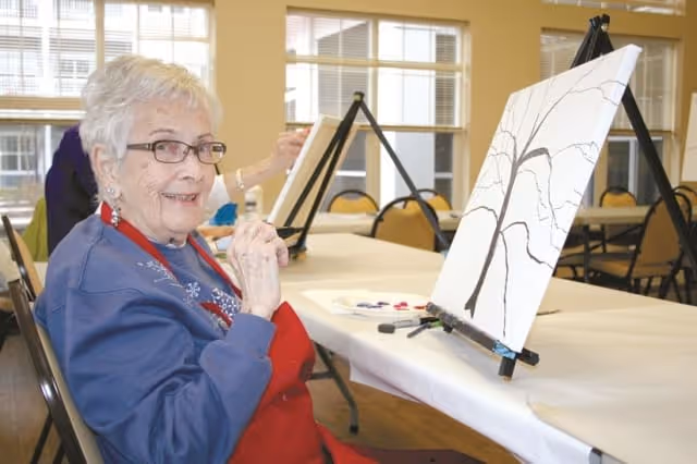 An elderly woman wearing glasses and a red apron is sitting at a table in a well-lit room, smiling at the camera while holding a paintbrush. In front of her is a canvas on an easel with a tree outline drawn on it, and paint supplies are on the table.