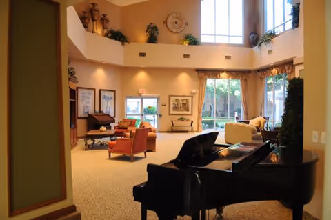Spacious and well-lit senior living community common area with a black grand piano in the foreground, comfortable seating including armchairs and sofas, large windows letting in natural light, decorative plants, and artwork on the walls.