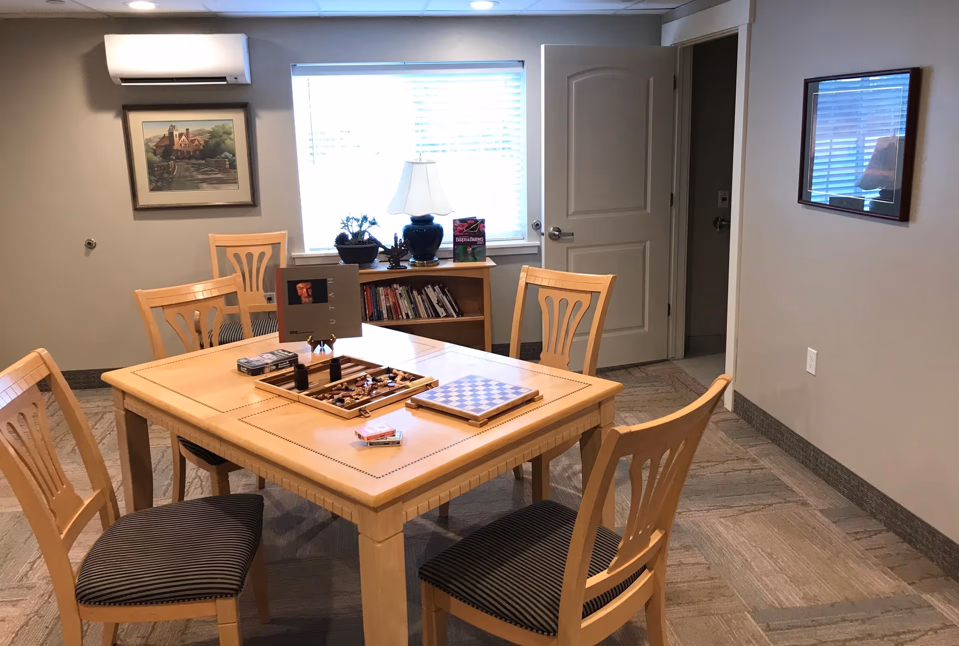 A light-wood table with four chairs arranged for board games in a communal room with a window, bookshelf, lamp, and an open door in the background.