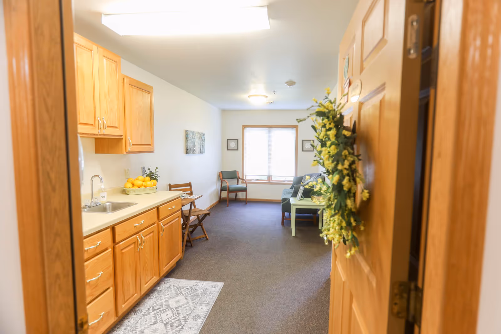 View through an open wooden door into a cozy senior living apartment featuring a kitchenette with wooden cabinets and a sink on the left, a small dining area with two chairs, and a living area with a sofa, coffee table, and armchair near a window with blinds. A yellow floral wreath hangs on the door.