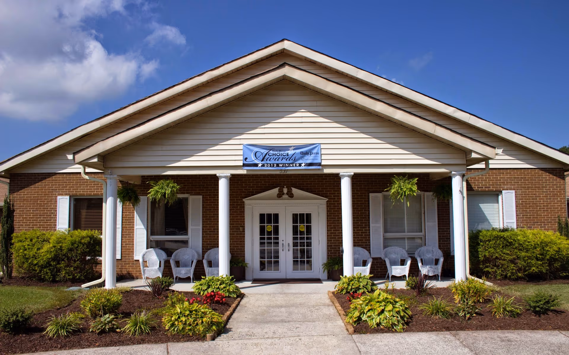 Front entrance of a single-story brick senior living building with a covered porch, white columns, chairs, hanging plants, and a landscaped walkway.