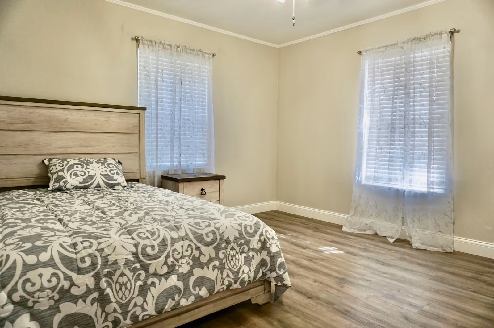 A bedroom with a wooden bed frame and a matching nightstand. The bed is made with a patterned gray and white comforter and a matching pillow. The room has two windows with sheer white curtains, light-colored walls, and wood flooring.