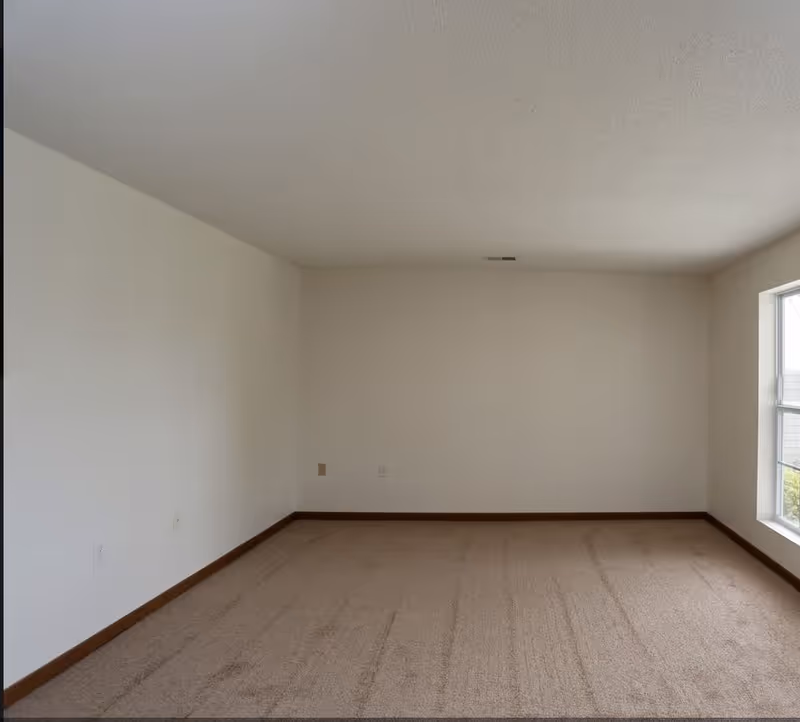 Empty room with beige carpet, white walls, and a large window on the right side letting in natural light.