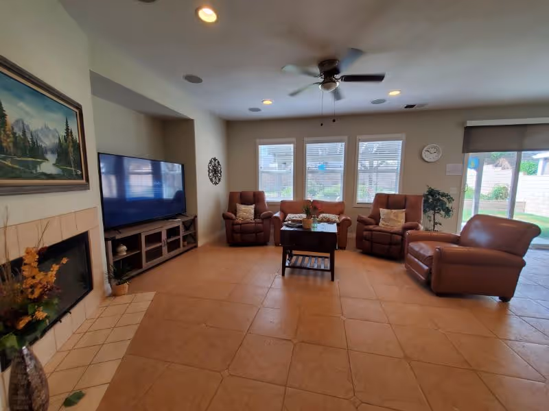 A spacious living room with beige tiled floor, a ceiling fan, and three large windows letting in natural light. The room features a flat-screen TV on a wooden stand, a fireplace with a floral arrangement, a painting above the fireplace, and four brown leather chairs arranged around a wooden coffee table. A sliding glass door leads to an outdoor area with a brick wall and greenery.