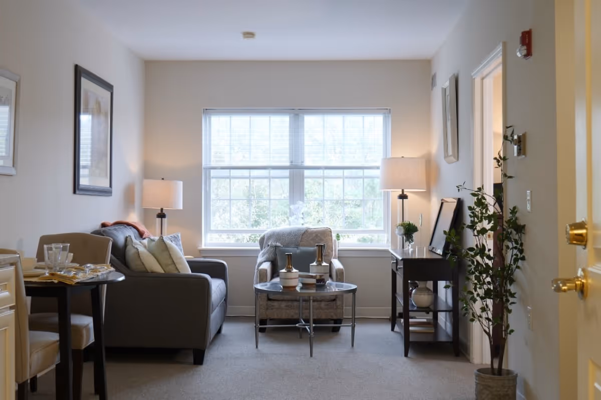 Well-lit living room with a sofa, armchair, coffee table, side tables with lamps, dining nook, and a large window.