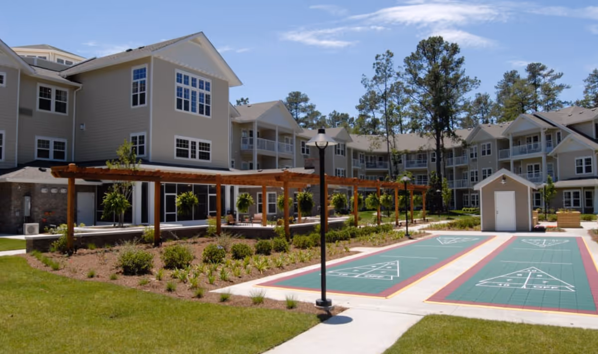 Outdoor area of Summerville Estates Gracious Retirement Living featuring a shuffleboard court, a wooden pergola with hanging plants, landscaped garden beds, and a multi-story residential building in the background under a partly cloudy sky.