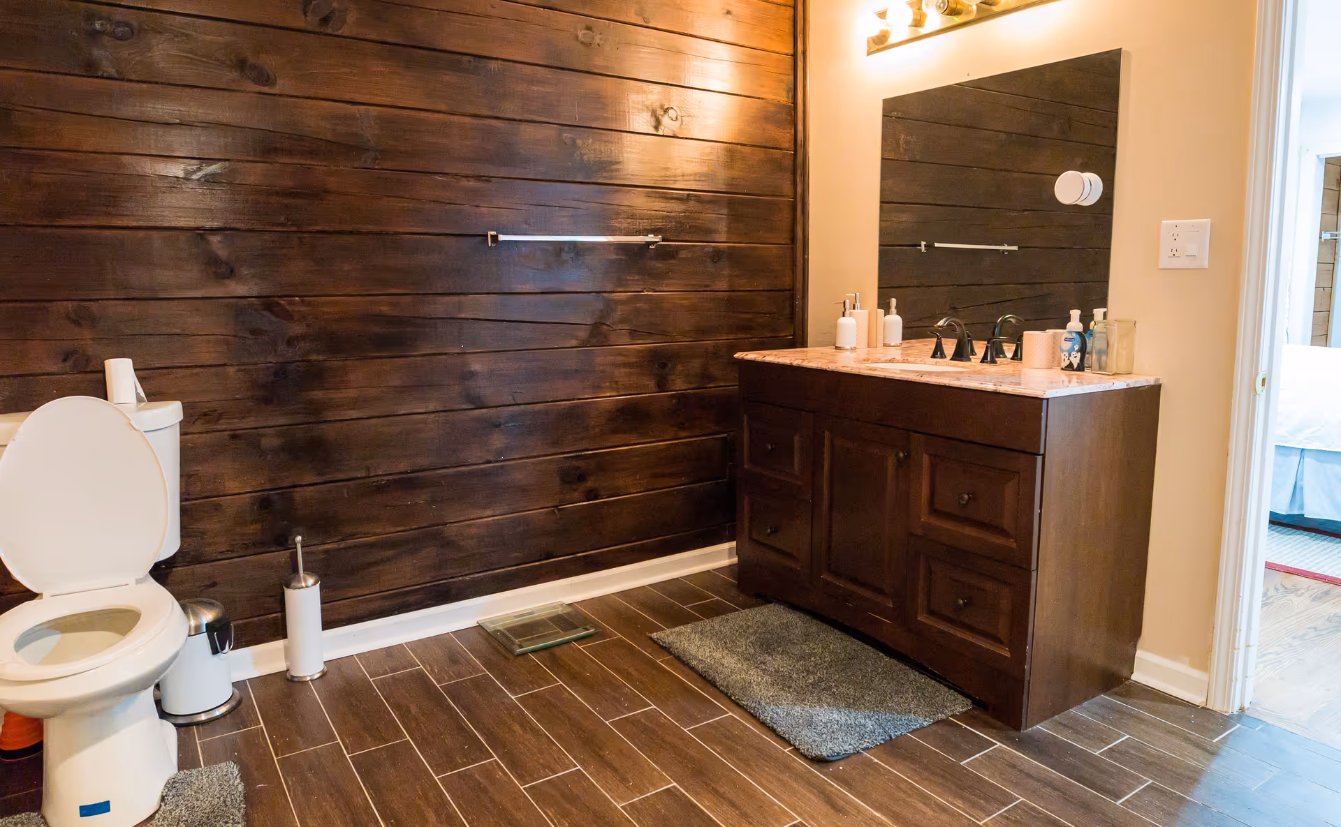 Bathroom with a white toilet, a wooden vanity with a marble countertop, a large mirror above the sink, and a dark wood paneled wall. The floor is tiled in a wood-like pattern and there are two gray rugs on the floor. A doorway leads to a bedroom with a bed visible.