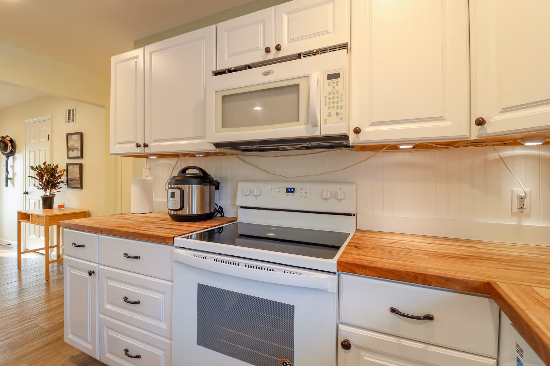 A bright kitchen with white cabinets and wooden countertops. There is a white electric stove with an oven and a white microwave mounted above it. A stainless steel electric pressure cooker and a paper towel roll are on the left countertop. The kitchen has under-cabinet lighting and a wooden floor. In the background, there is a small wooden table with a potted plant and some wall decorations near a white door.