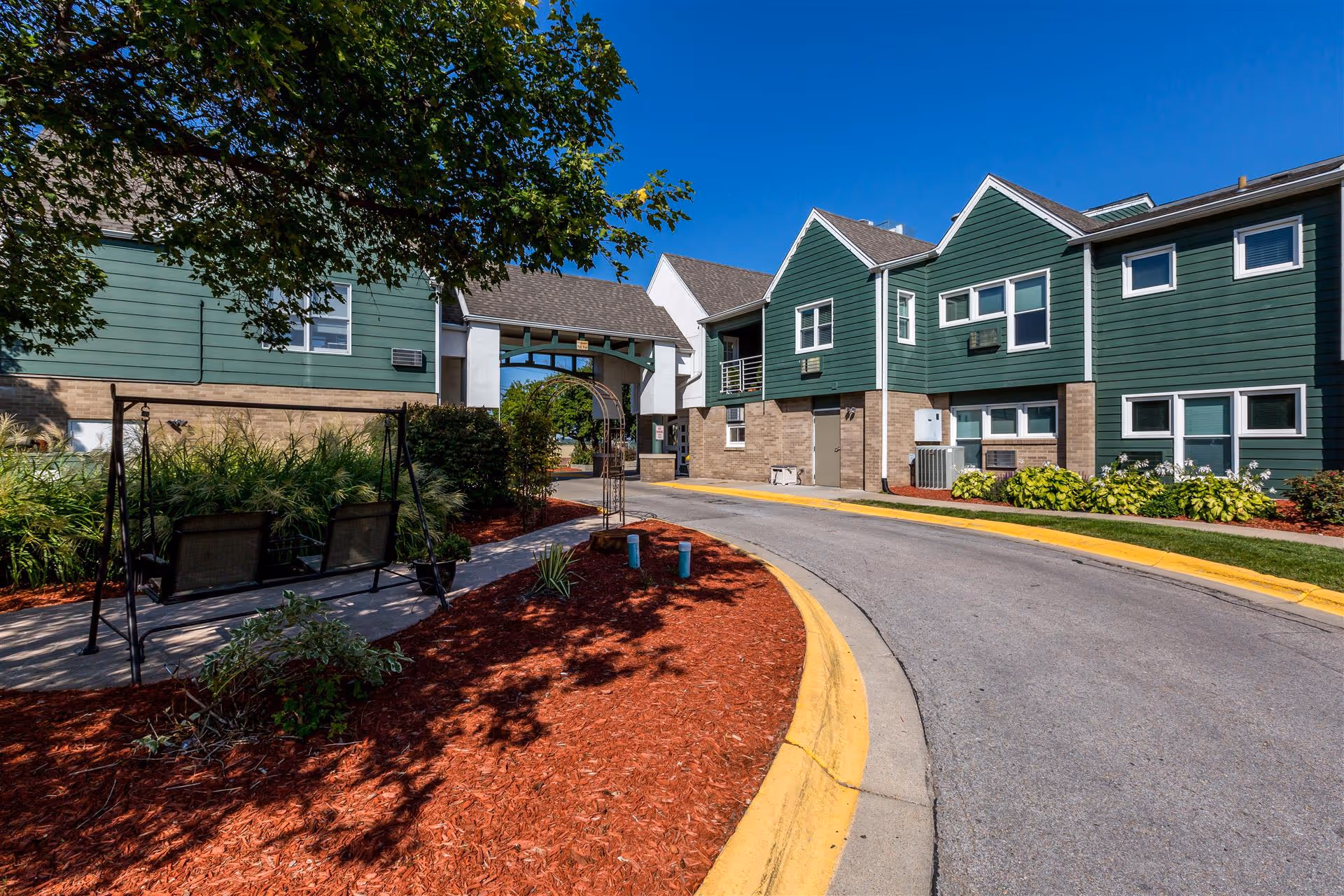 Outdoor view of a senior living facility with green and beige buildings, a curved driveway, landscaped garden beds with red mulch, and a black metal swing bench under a tree.