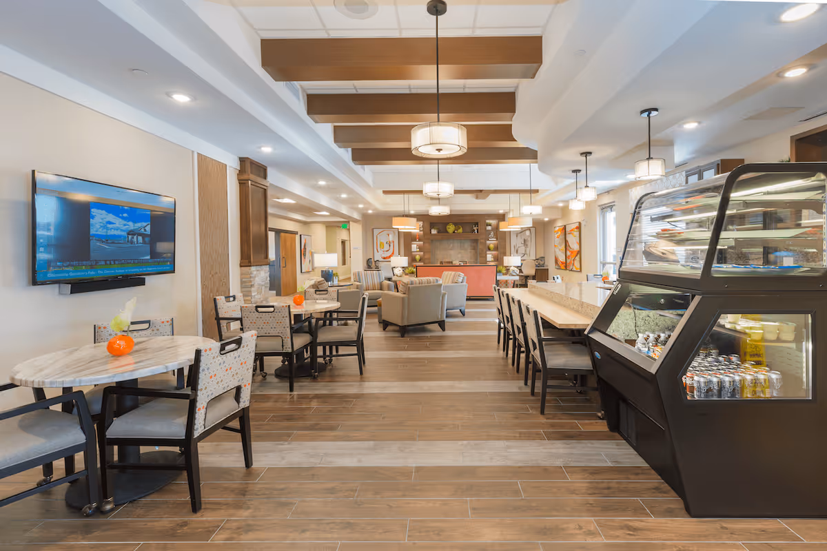 A spacious and well-lit common area in a senior living facility featuring multiple round tables with chairs, a long counter with high stools, a glass display case with beverages and snacks, comfortable armchairs arranged near a fireplace, and modern ceiling lights with wooden beams.