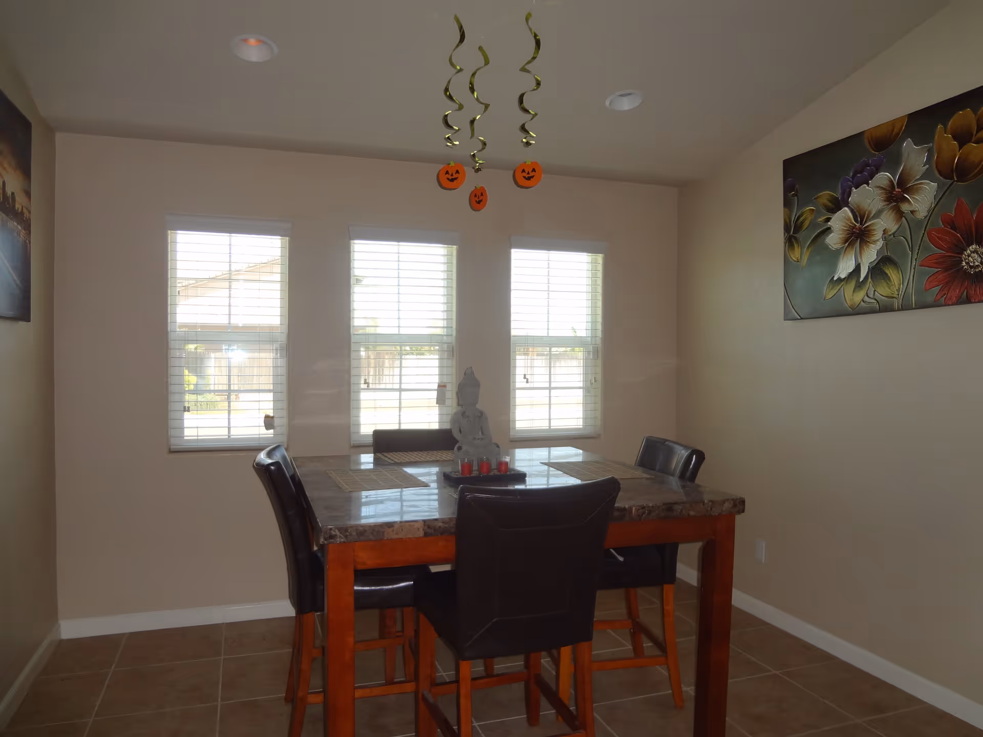 A dining area with a square table featuring a marble-like top and four dark leather chairs. Three windows with white blinds are on the wall behind the table. Hanging from the ceiling are three spiral decorations with orange pumpkin faces. There are two paintings on the walls, one with a cityscape and the other with colorful flowers.