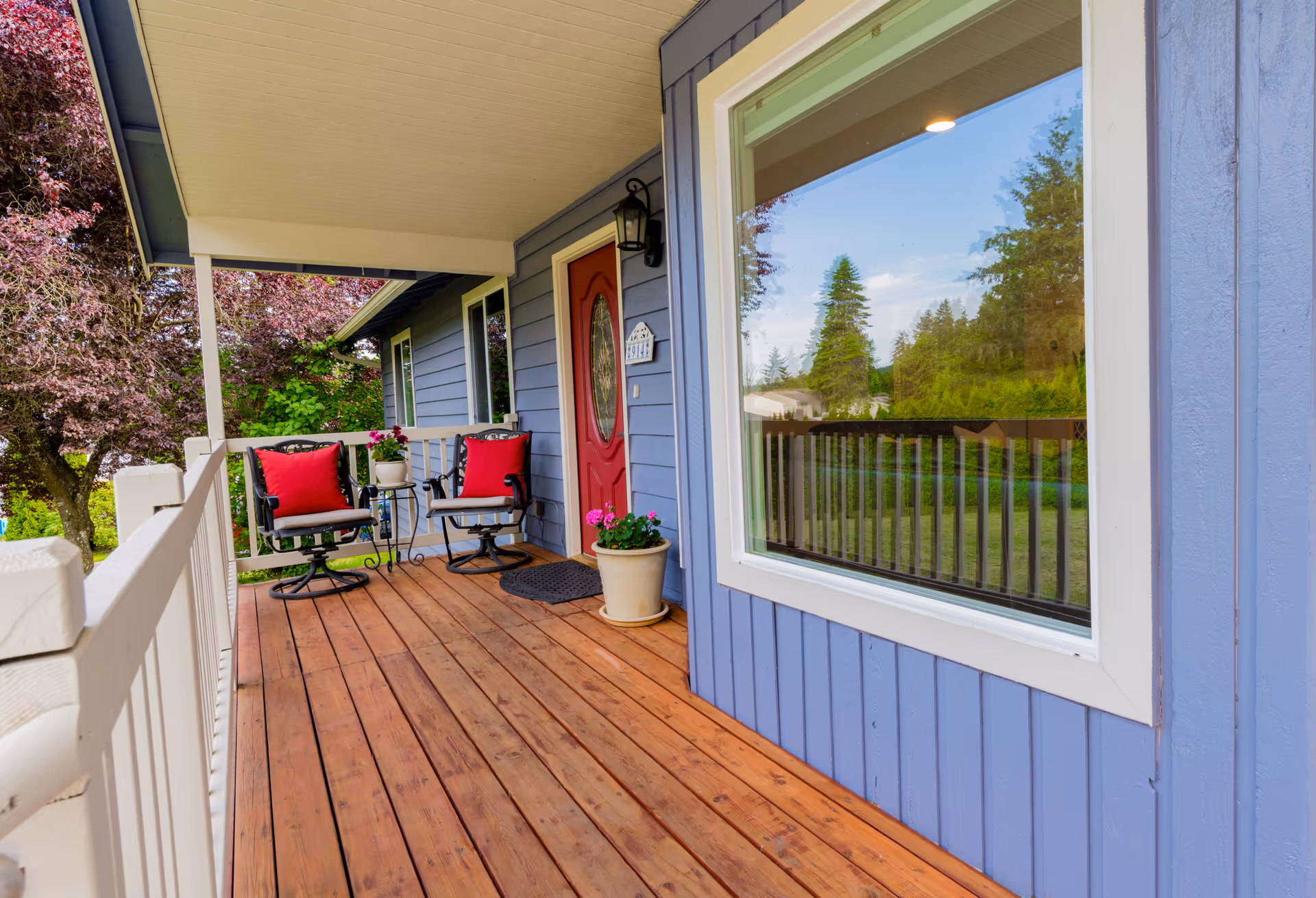 Covered wooden porch with two black metal chairs with red cushions and a small round table with potted plants. The porch has a red front door, a large window with white trim, and overlooks a green yard with trees.