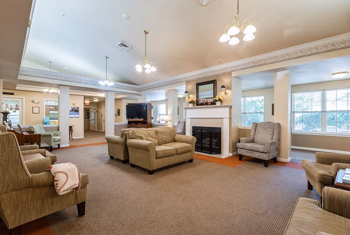 A spacious living room area in a senior living facility with beige armchairs and sofas arranged around a fireplace. The room has large windows letting in natural light, a TV on a stand, and decorative lighting fixtures hanging from the ceiling. The walls are painted in neutral tones with crown molding along the ceiling edges.
