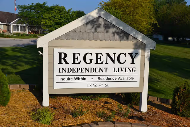 Outdoor sign for Regency Independent Living with text Inquire Within, Residence Available, and address 418 W. 4th St. The sign is mounted on a small structure with a shingled roof, surrounded by grass and landscaping with trees and a building in the background.