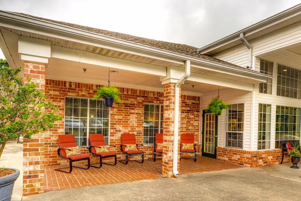 Covered outdoor patio area at Bridgewood Gardens with five red cushioned chairs arranged against a brick wall, two hanging green plants, and large windows and a door leading inside the building.