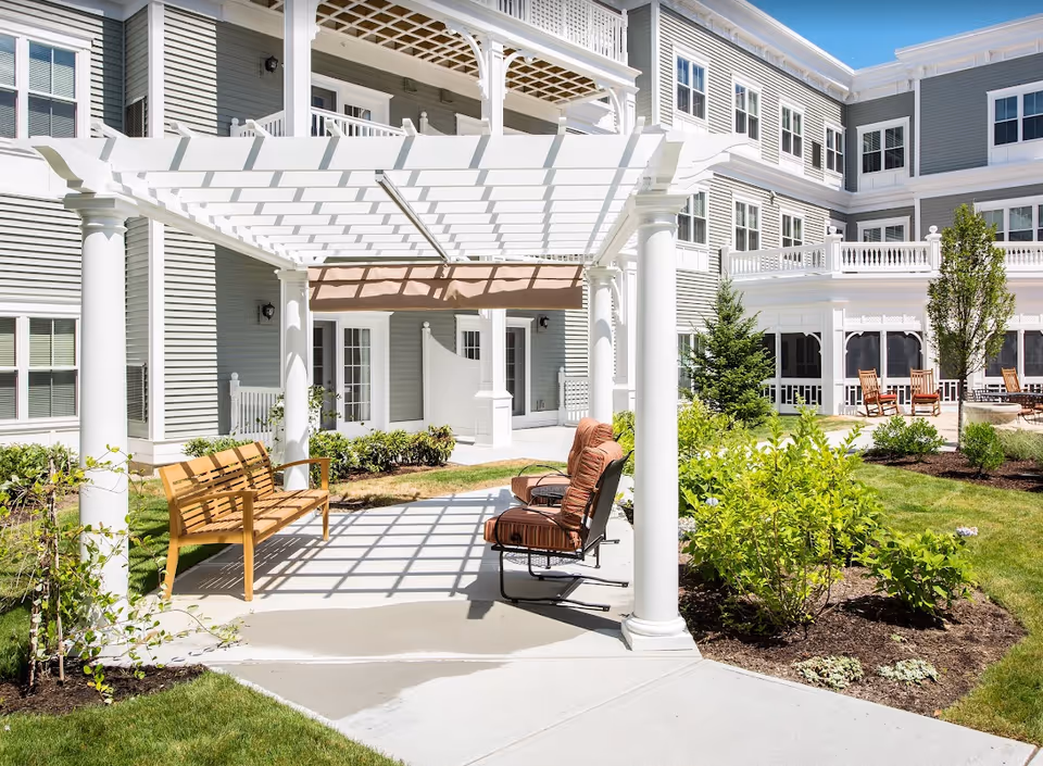 Sunny courtyard with a white pergola, benches and cushioned chairs in front of a multi-story senior living building.
