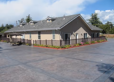 Single-story beige senior living building with a fenced patio, landscaping, and a large paved courtyard under a partly cloudy sky.