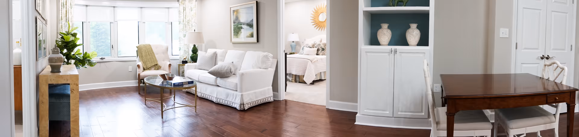 A bright and spacious interior living area with wooden flooring, a white sofa, a glass coffee table, and a chair near large windows with white blinds. There is a small console table with a plant and decorative items on the left, a built-in white cabinet with vases on the right, and a wooden dining table with chairs. In the background, a bedroom with a bed and bedside lamps is visible through an open doorway.