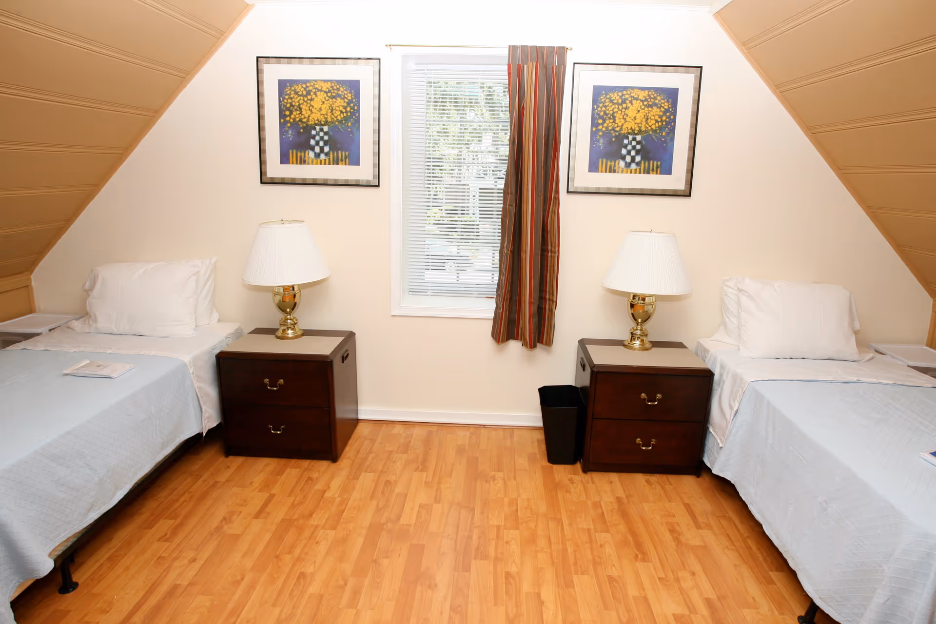 Two twin beds with nightstands and lamps flanking a central window in a simple bedroom with hardwood floors.