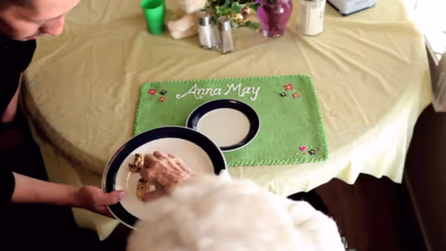 A person hands a plate of food to an elderly person at a round table with a green placemat embroidered 'Anna May'.
