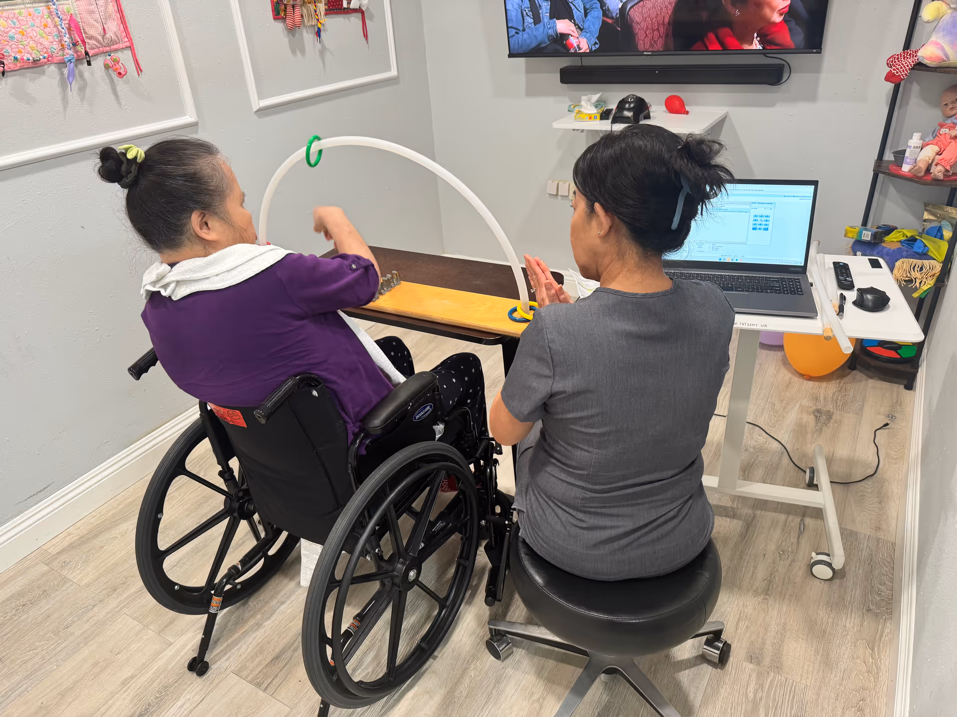 Two women in a therapy or activity room; one woman in a wheelchair wearing a purple top is engaging with a ring toss game on a wooden board, while the other woman in gray scrubs sits on a stool beside her, clapping. A laptop and TV screen are visible in the background along with shelves holding toys and supplies.