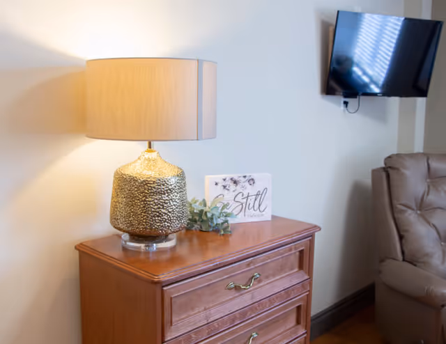 A cozy corner of a living room featuring a wooden chest of drawers with a textured gold lamp, a small decorative plant, and a sign that reads 'Be Still'. A wall-mounted flat-screen TV and part of a cushioned armchair are visible in the background.