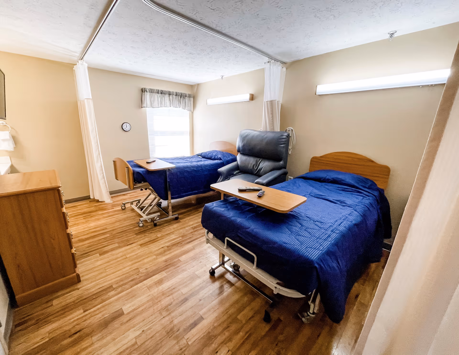 Shared resident bedroom with two hospital-style beds, a recliner, overbed tables, and wood-look flooring.