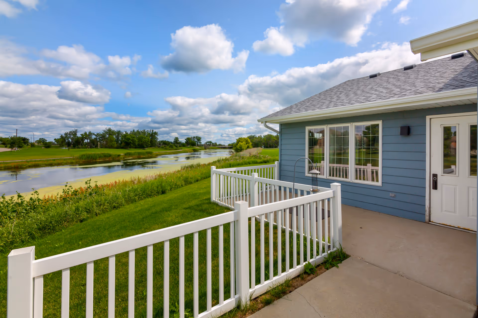 Outdoor patio area of a senior living facility with a white railing, concrete floor, and blue exterior walls. The patio overlooks a grassy area and a calm river with trees and a partly cloudy sky in the background.