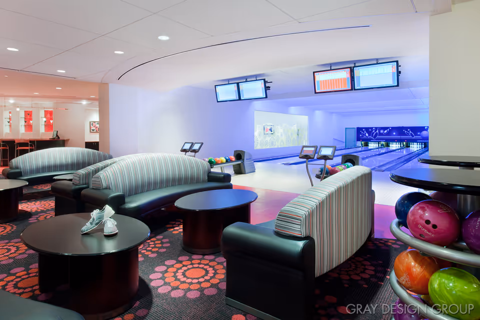 Modern indoor bowling alley lounge with striped sofas, round tables, and visible lanes and colorful bowling balls.