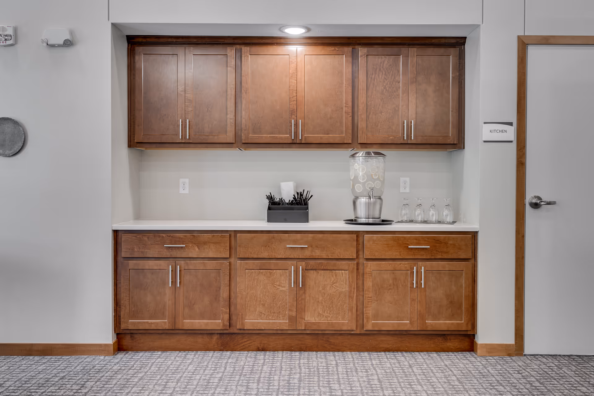 Built-in wooden kitchenette with upper and lower cabinets, a countertop water dispenser, glasses and utensils.
