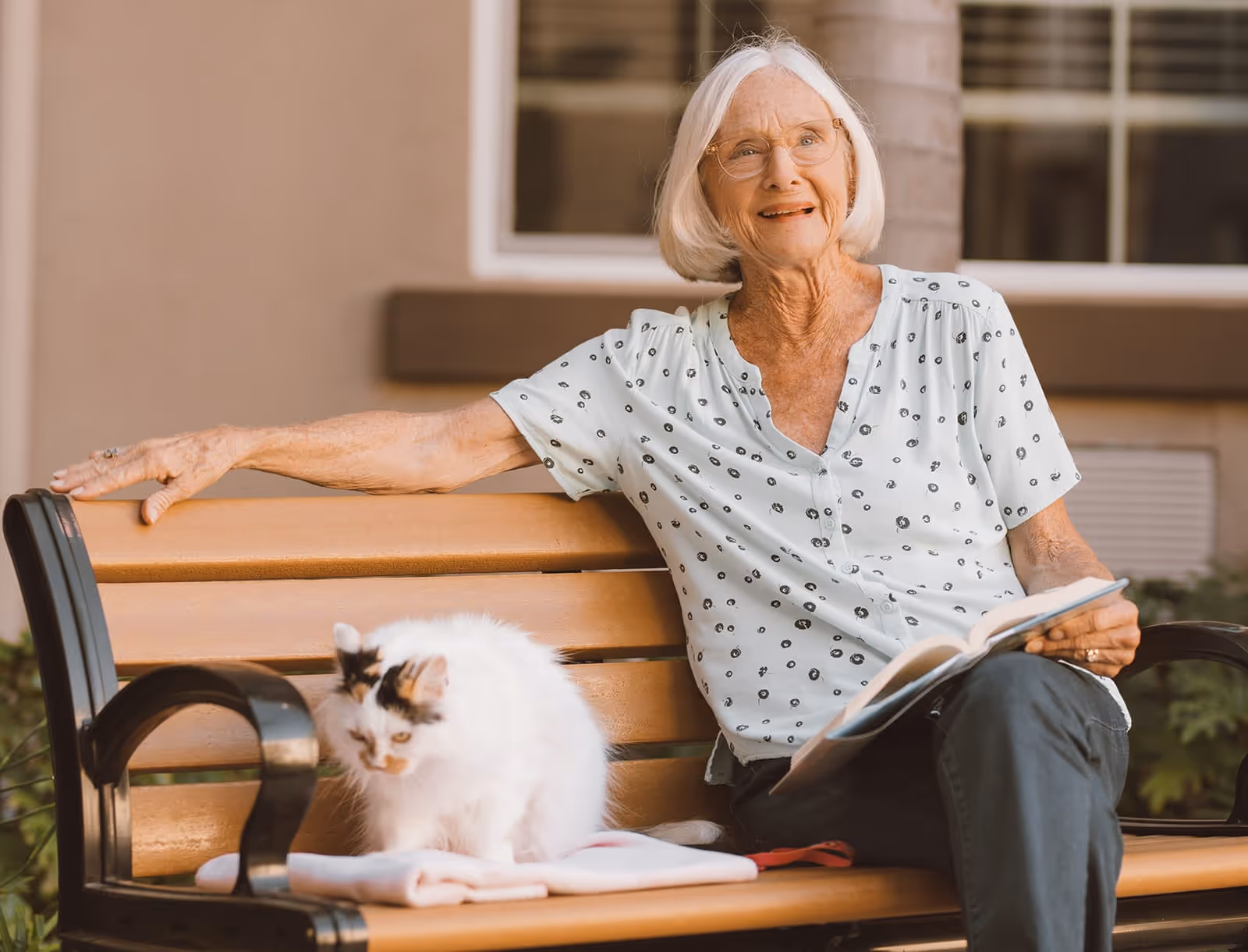 An elderly woman with white hair and glasses sitting on a wooden bench outdoors, smiling and holding an open book. A white cat with black and orange spots is sitting beside her on the bench on a folded blanket.