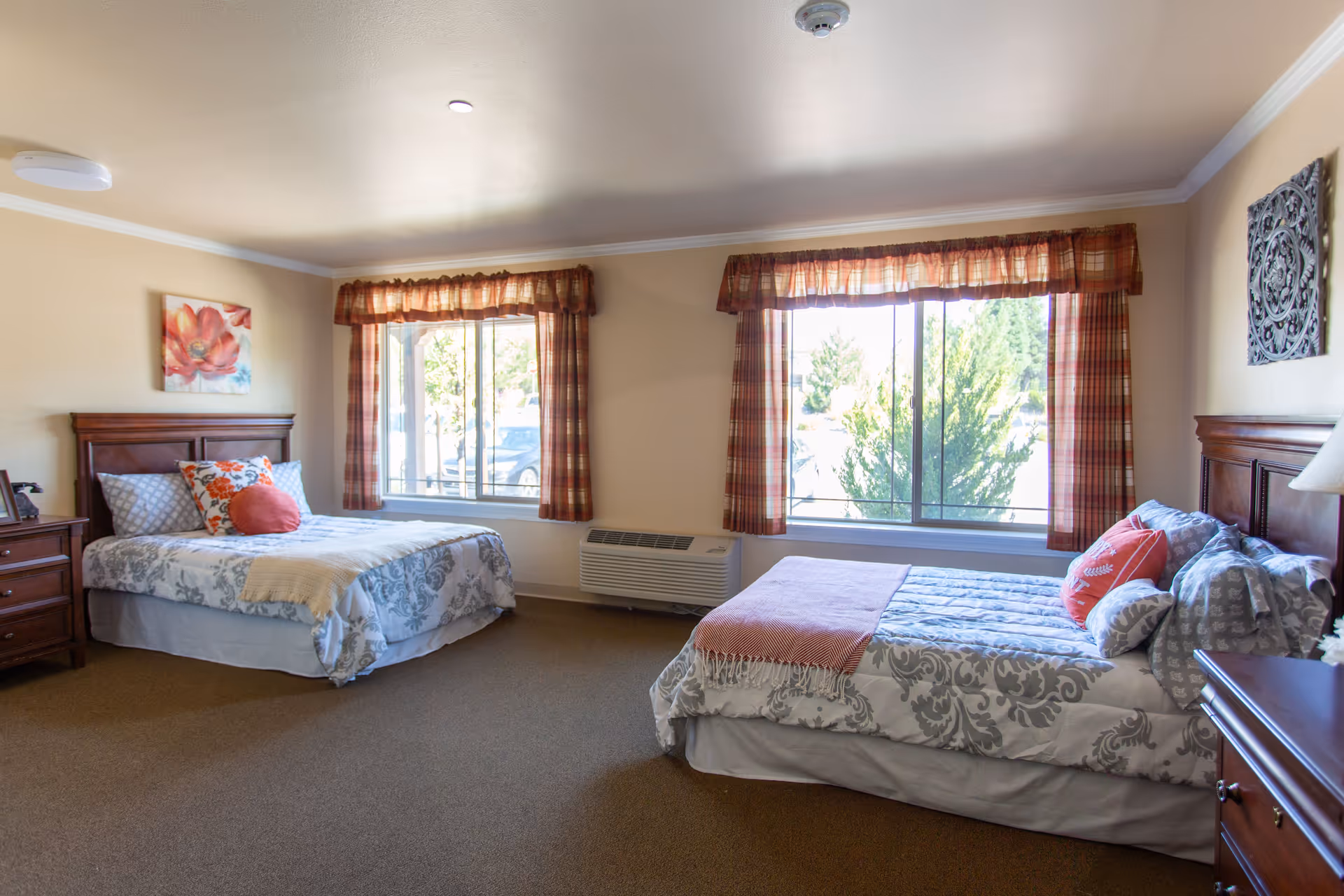 A bright bedroom with two beds, each with patterned bedding and decorative pillows. The room has two large windows with plaid curtains letting in natural light. There are wooden nightstands beside each bed, a floral painting on the wall above one bed, and a decorative wall piece above the other. The carpet is brown and the walls are painted a light beige color.