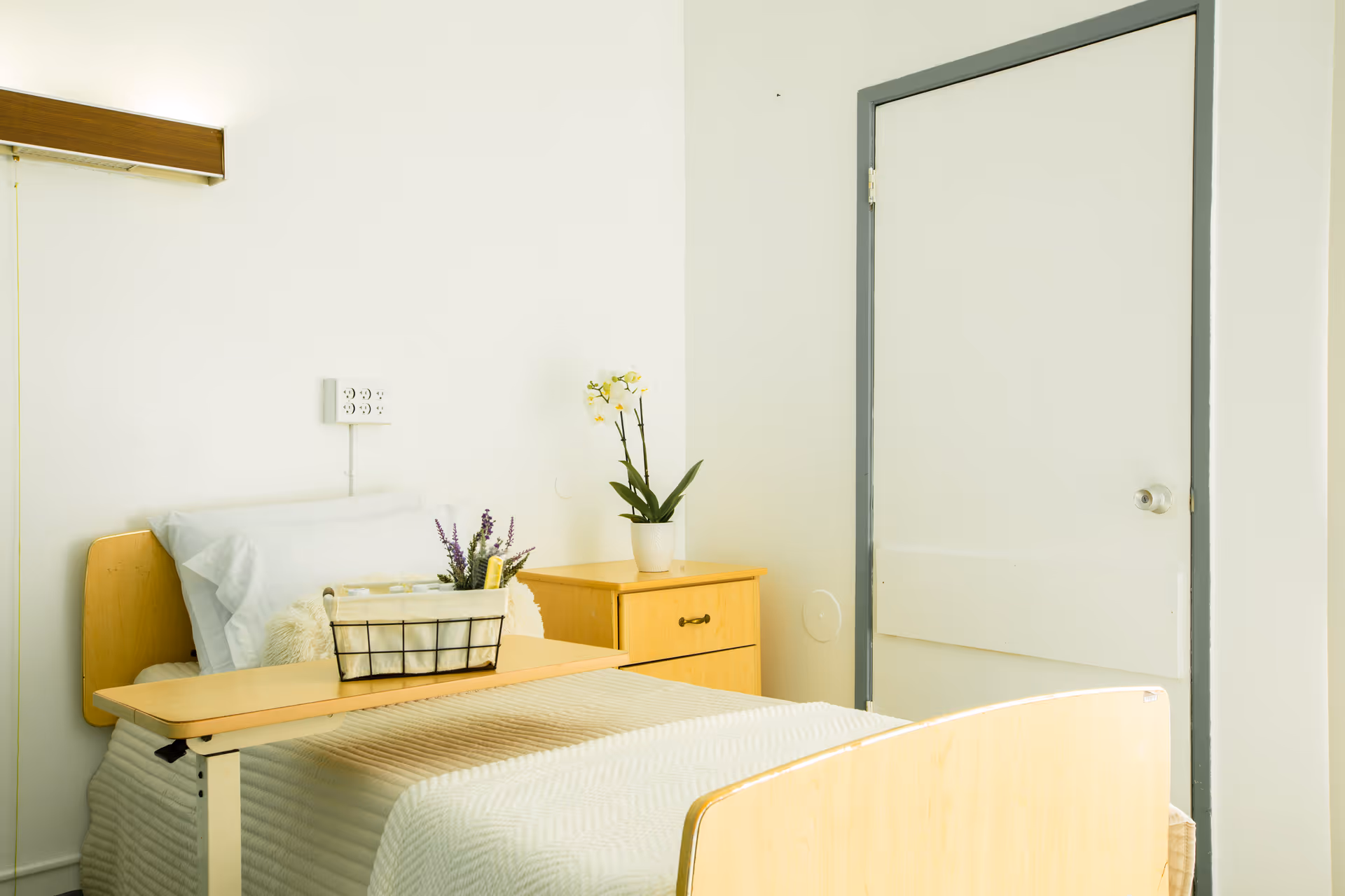 A simple, clean bedroom in a healthcare facility with a single bed covered in a beige blanket, a wooden bedside table with a potted orchid plant, and a small table over the bed holding a basket with toiletries and flowers. The walls are white, and there is a closed door with a gray frame.