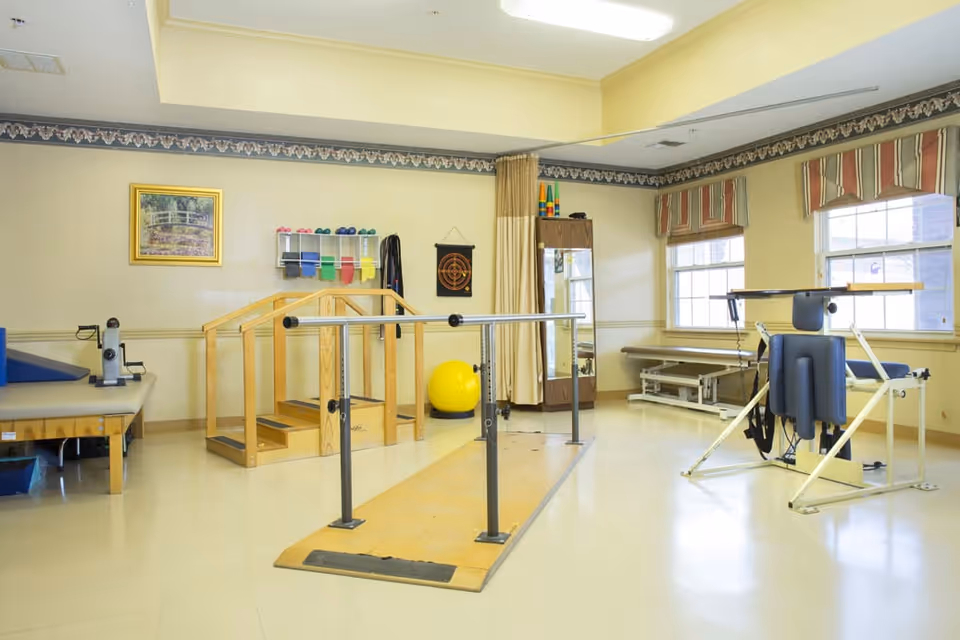 A physical therapy room with parallel bars for walking exercises, a small wooden staircase for step training, various exercise equipment including a yellow exercise ball, therapy tables, and a large mirror. The room has beige walls, windows with striped valances, and a framed picture on the wall.
