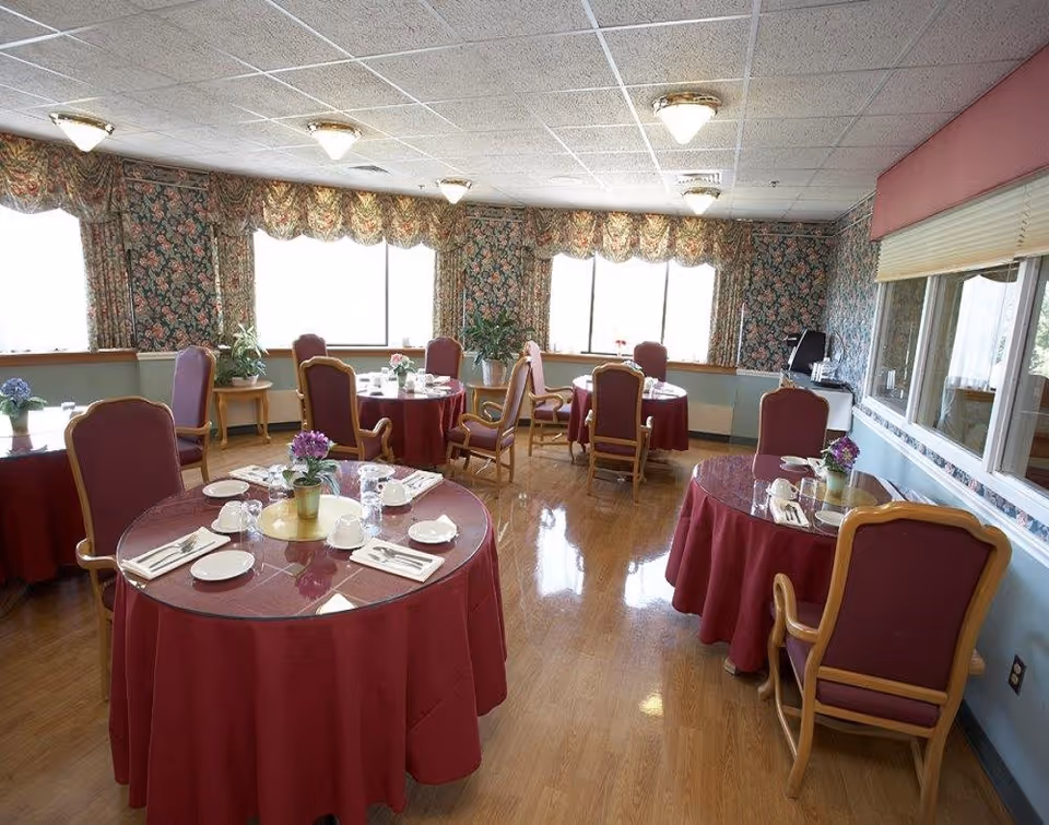Dining room with round tables dressed in red tablecloths, place settings, upholstered chairs, and floral curtains and wallpaper.