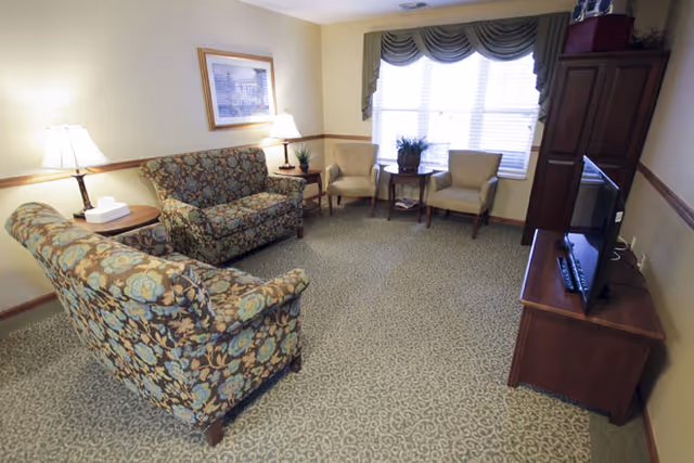 A cozy living room with two floral patterned sofas, two beige armchairs, a wooden TV stand with a flat-screen television, a side table with a lamp and tissue box, and a window with blinds and a valance. The room has carpeted flooring and soft lighting.