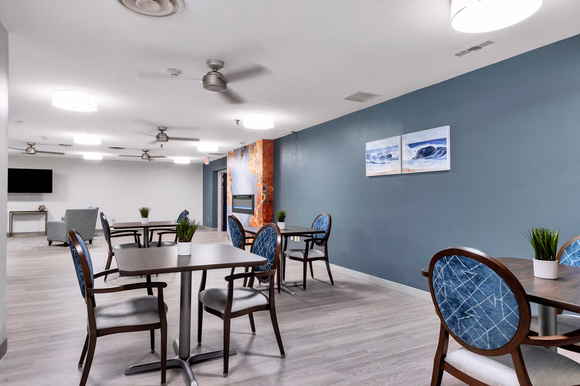 A communal dining and lounge area with several tables and upholstered chairs against a blue accent wall and a seating area with a TV.
