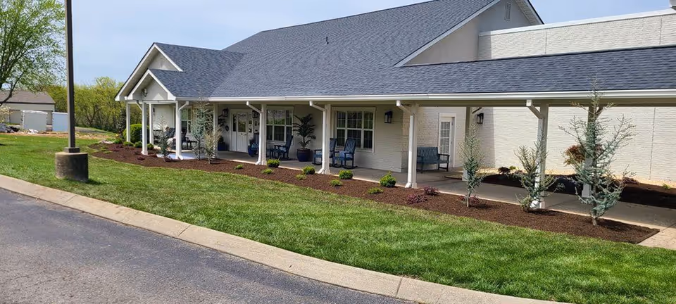 Exterior view of a single-story building with a covered porch supported by white columns. The porch has several chairs and potted plants. The building is surrounded by a well-maintained lawn and landscaped garden beds with small shrubs and young trees. A street lamp is visible on the left side near the curb.