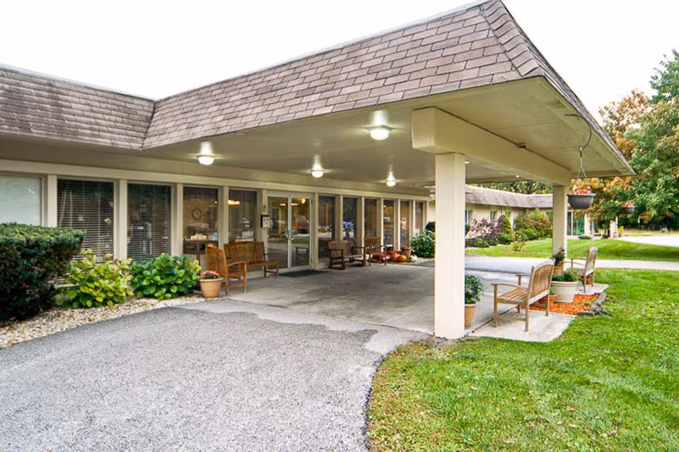Covered entrance and drive-up drop-off area of a senior living facility with benches, potted plants, and glass doors.