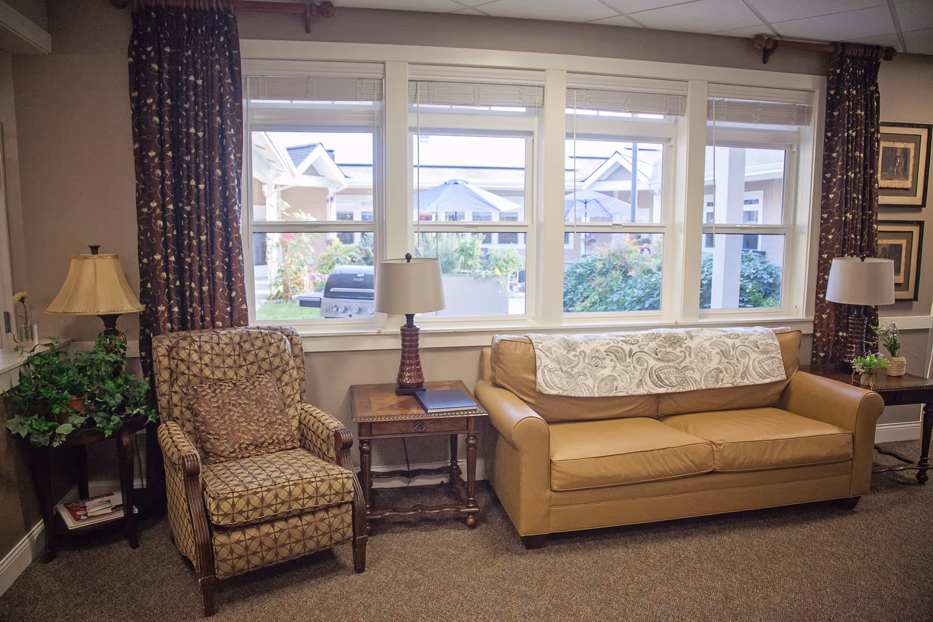 A cozy living room area with a patterned armchair and a tan leather sofa. Between them is a wooden side table with a lamp and a book. There are large windows behind the furniture showing an outdoor view of a courtyard with greenery and buildings. The room has carpeted flooring, curtains on either side of the windows, and additional side tables with lamps and plants.