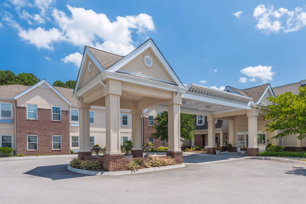 Exterior view of Park Place of West Knoxville senior living facility entrance with a covered drop-off area, beige and brick building facade, and clear blue sky with scattered clouds.
