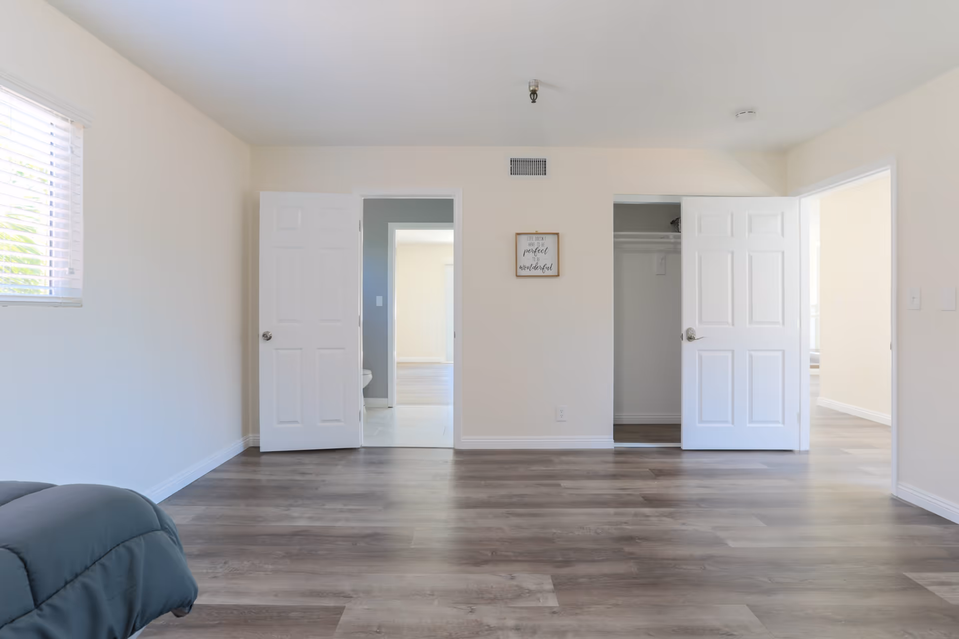 Empty room with light-colored walls and wood flooring, featuring three open doors leading to a bathroom, a closet, and another room. A small framed sign is hung on the wall between the doors. A window with blinds is on the left side, and part of a bed with a dark comforter is visible in the bottom left corner.