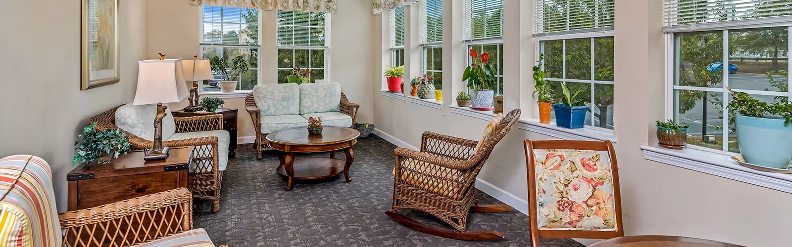 Sunlit common sitting area with wicker chairs, floral-upholstered seating, wooden tables and potted plants along a row of windows.