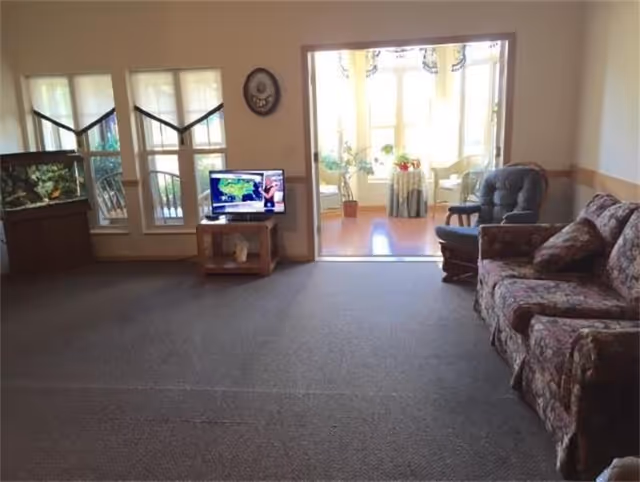 A living room with a floral patterned couch on the right, a cushioned rocking chair next to it, a small TV on a wooden stand showing a weather forecast, and a large fish tank on the left near windows. The room opens into a sunlit area with large windows, plants, and a small table with chairs.