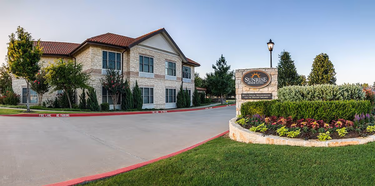 Exterior front view of the Sunrise senior living building with a driveway, landscaped flowerbed, and an entrance sign.